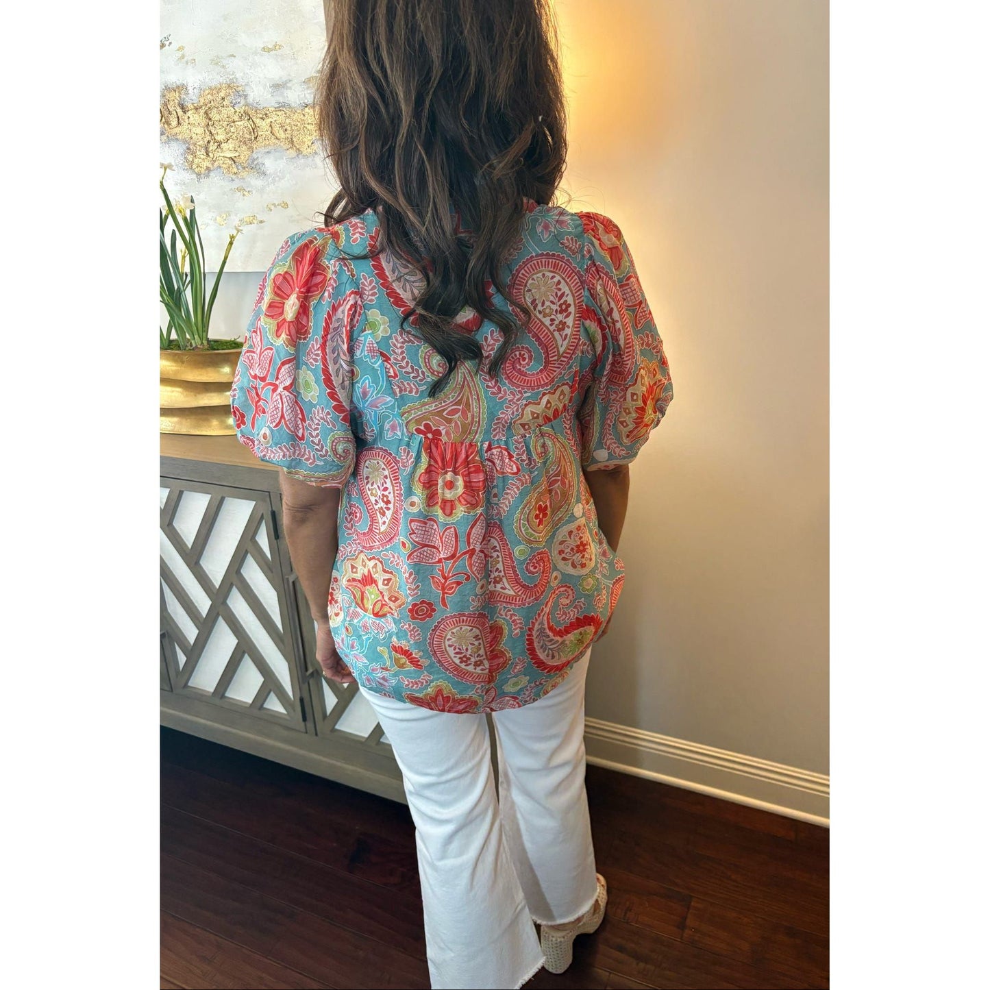 Person wearing a colorful paisley shirt and white pants in a room with a decorative wall and plant.