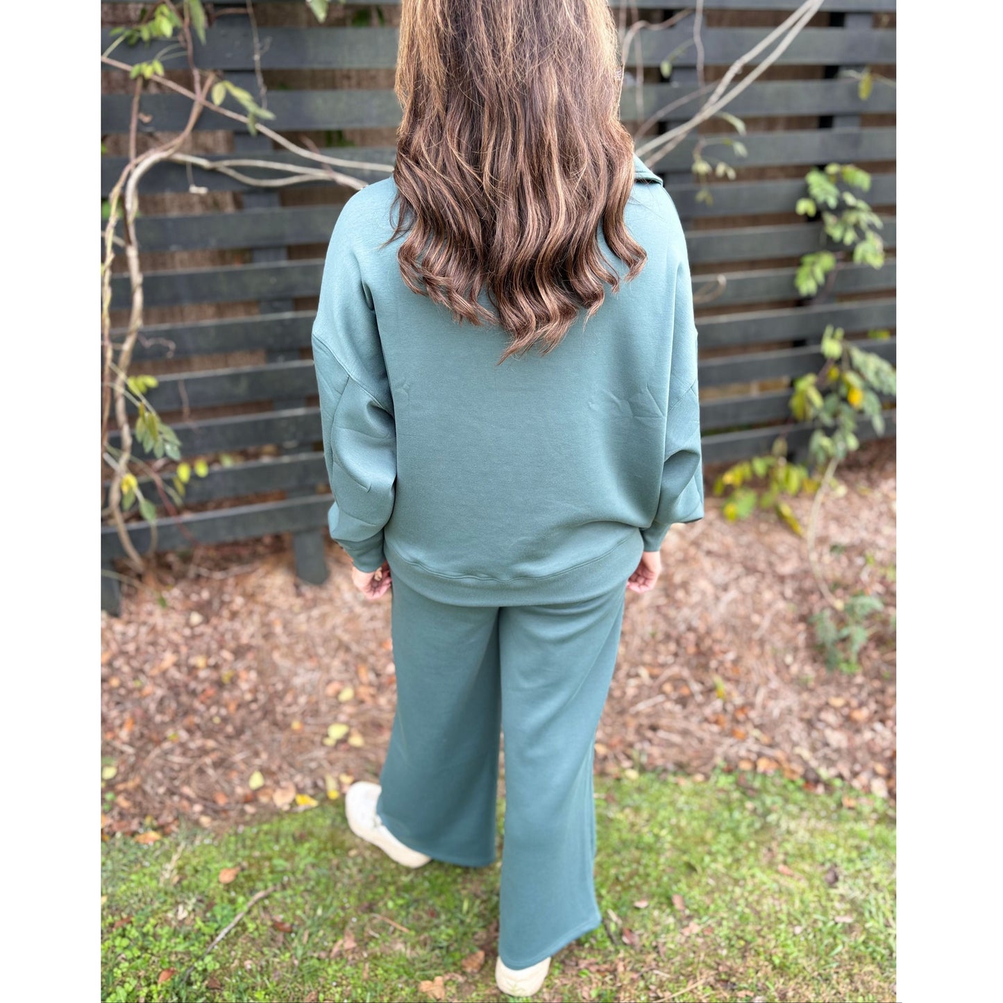 Person wearing a teal tracksuit standing outdoors with a wooden fence and grass in the background