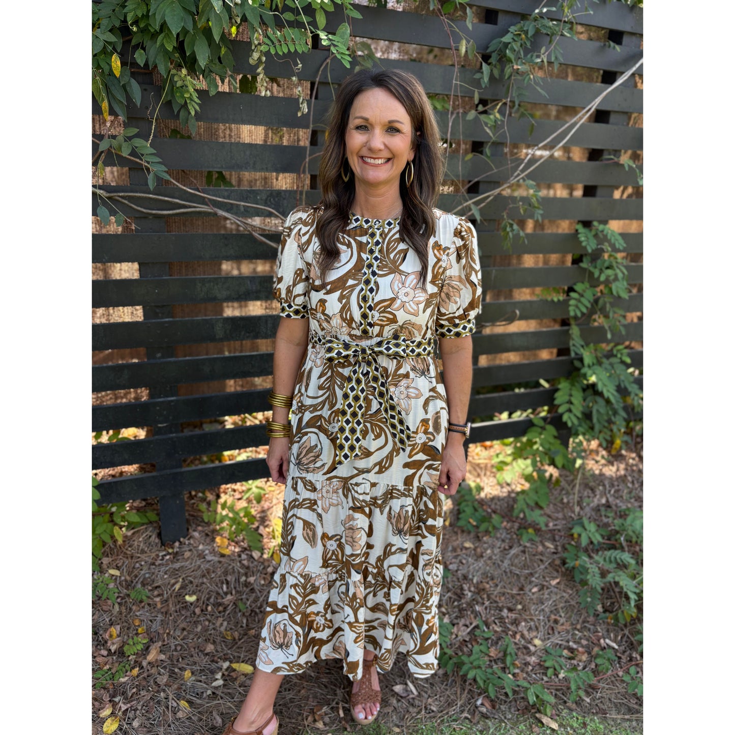 Woman wearing a patterned dress standing in front of a wooden fence with greenery.
