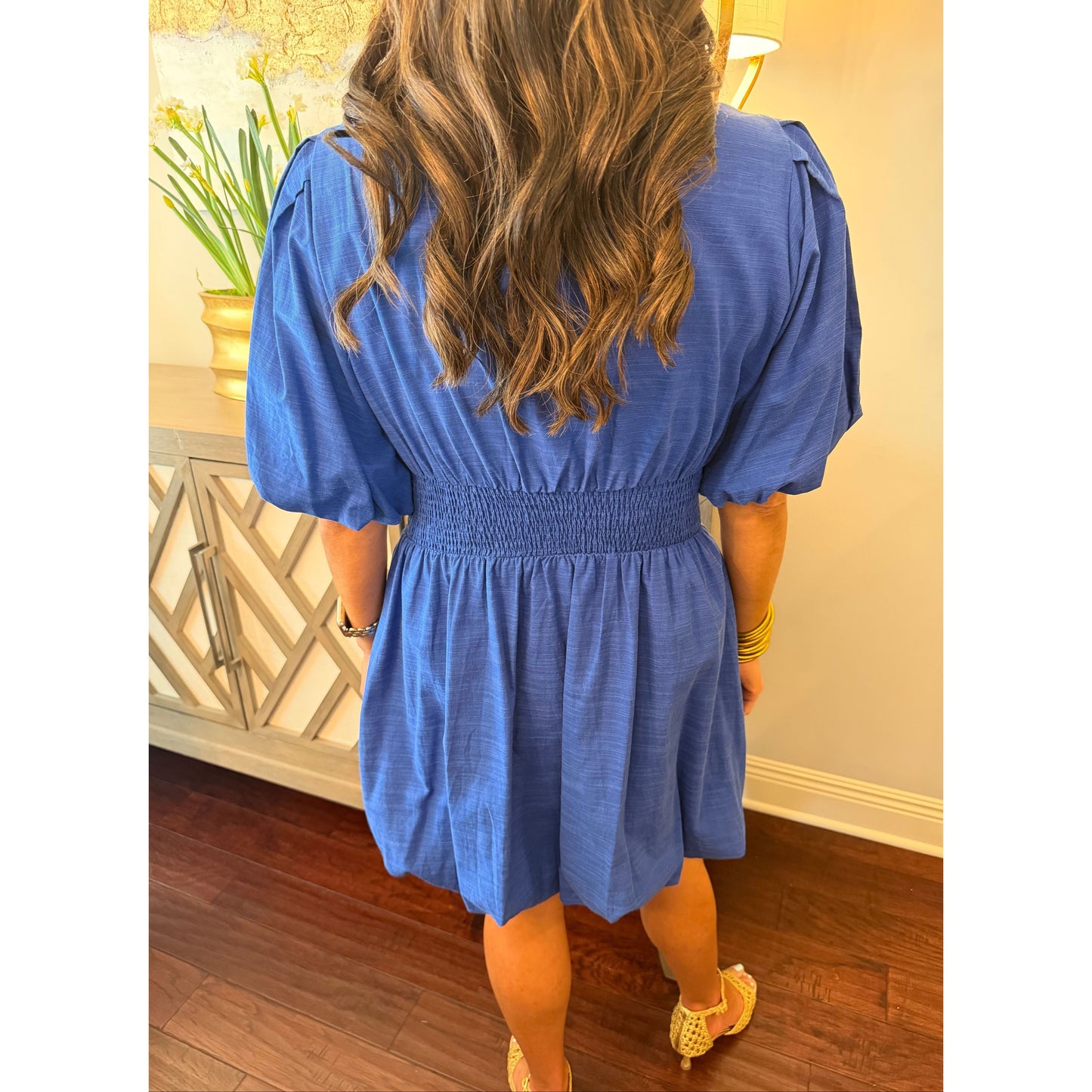 Person wearing a blue dress in an indoor setting with wooden flooring and a decorative plant.