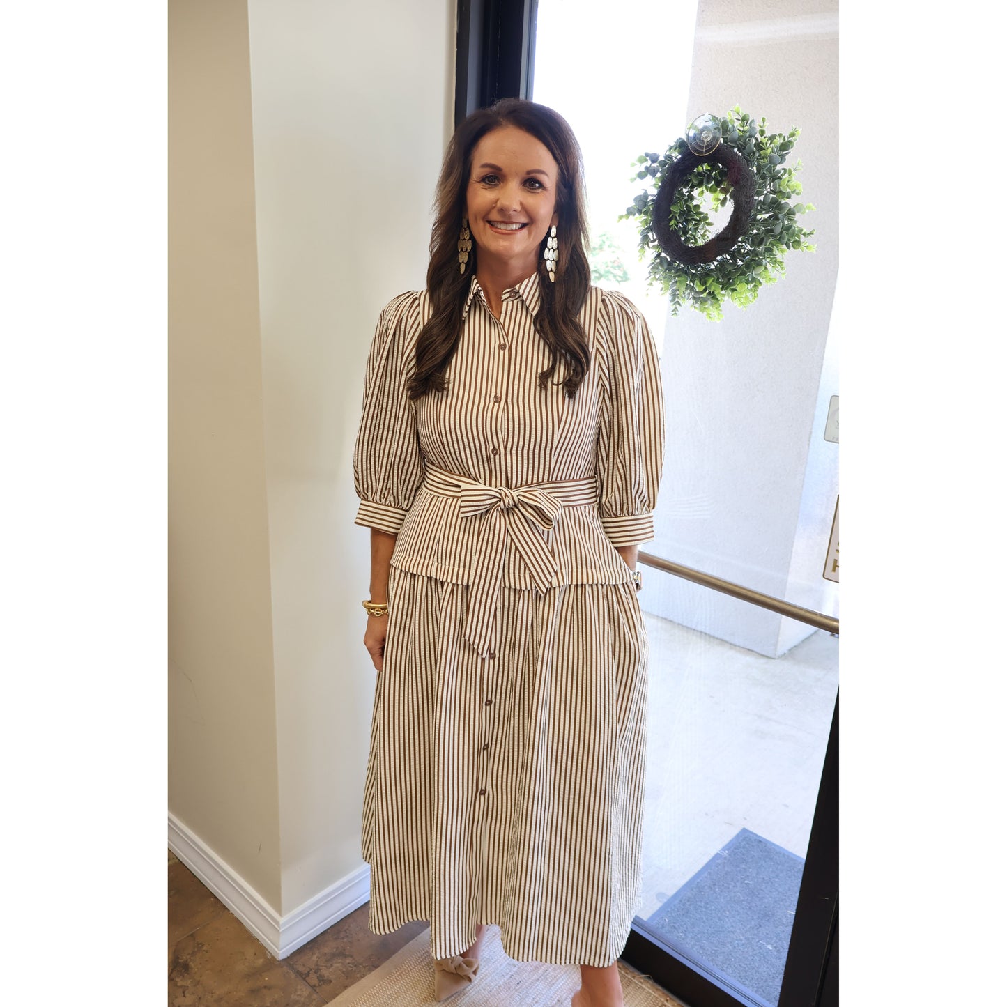 A woman wearing a beige and brown vertically striped midi shirt dress with puff sleeves, a button-down front, and a matching tie waist belt, standing in an indoor setting.