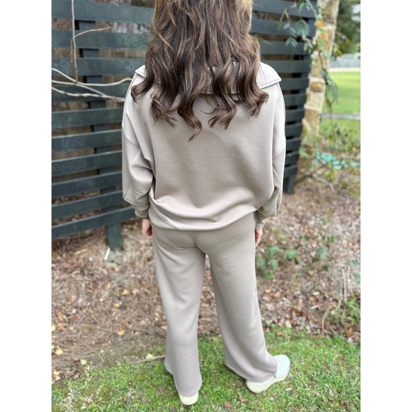 Person wearing a beige tracksuit standing outdoors with a wooden fence and grass in the background