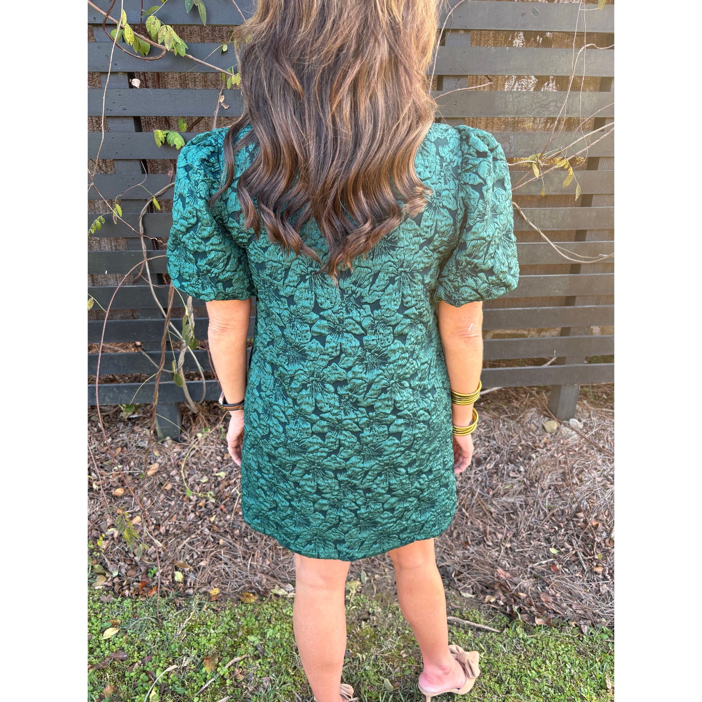 Person wearing a green textured dress standing outdoors with a wooden fence and grass in the background.