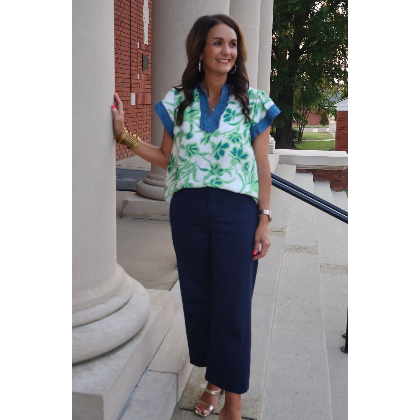Woman in a floral top and navy pants standing outdoors on a stone path.