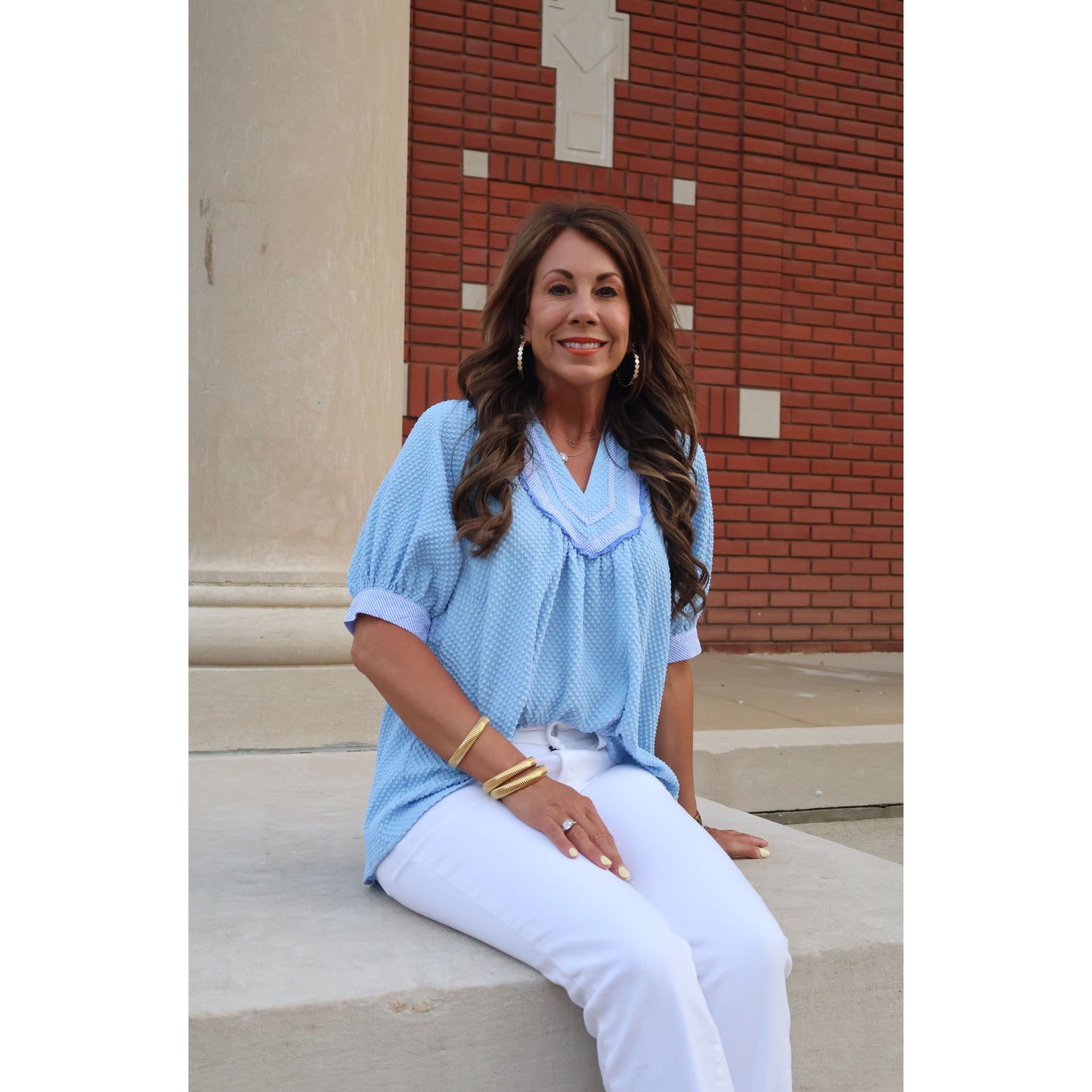 Woman in a light blue top and white pants standing against a brick wall.