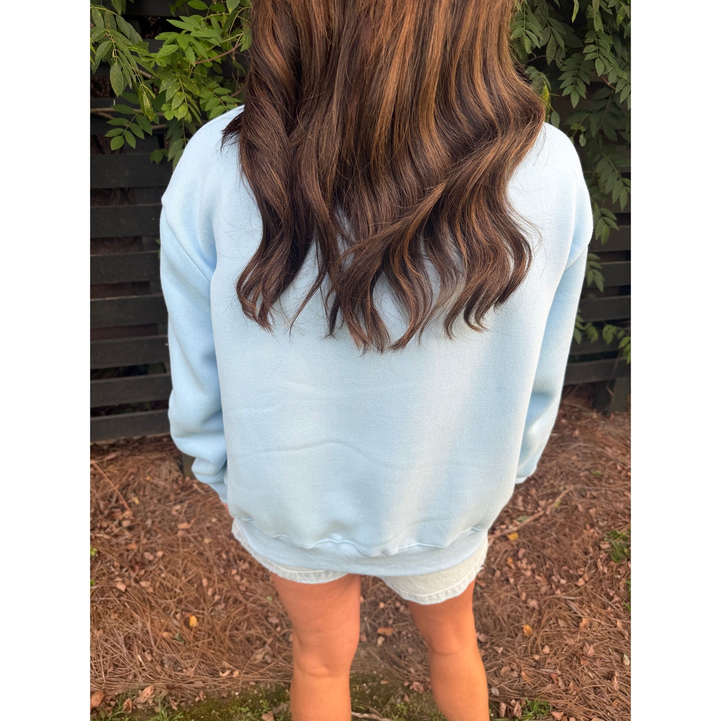 A person viewed from behind wearing a light blue sweatshirt and white denim shorts, standing outdoors on a mulch ground with green foliage and a dark fence in the background.