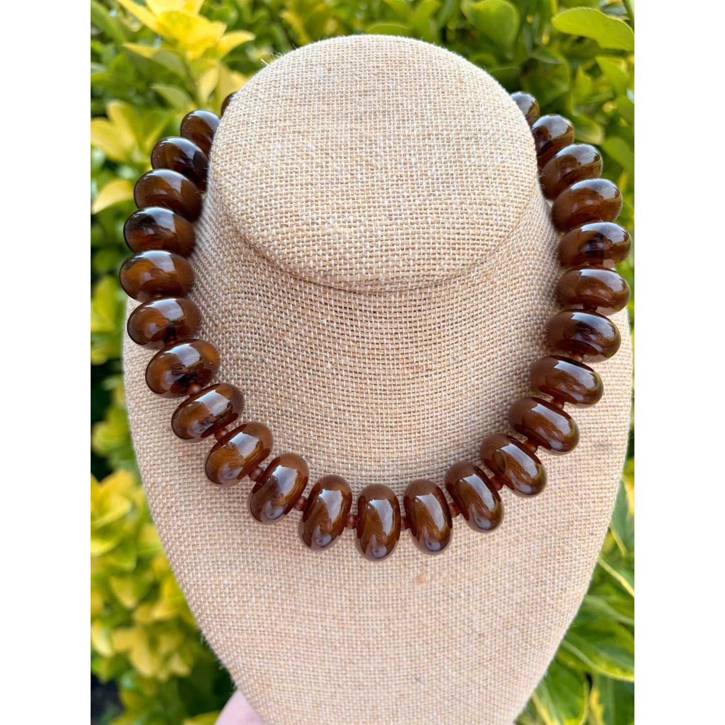 A dark brown glossy beaded necklace displayed on a beige fabric-covered jewelry bust, with blurred green foliage in the background.