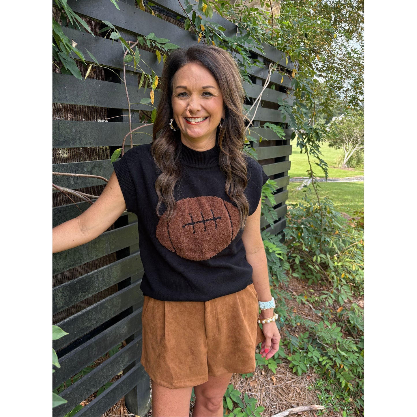 Woman wearing a black t-shirt with a brown football design and brown shorts, standing outdoors.