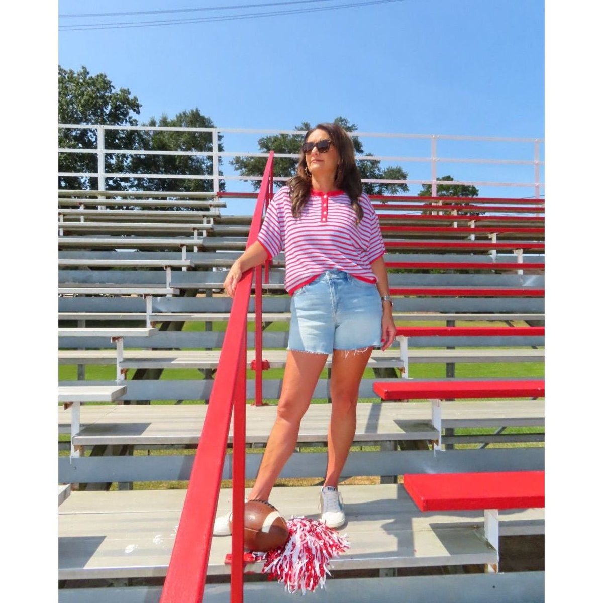 A woman stands on red and gray metal bleachers outdoors, wearing a pink and white striped short-sleeve polo shirt, denim shorts, white sneakers, and sunglasses. She holds a football and a red-and-white pom-pom.