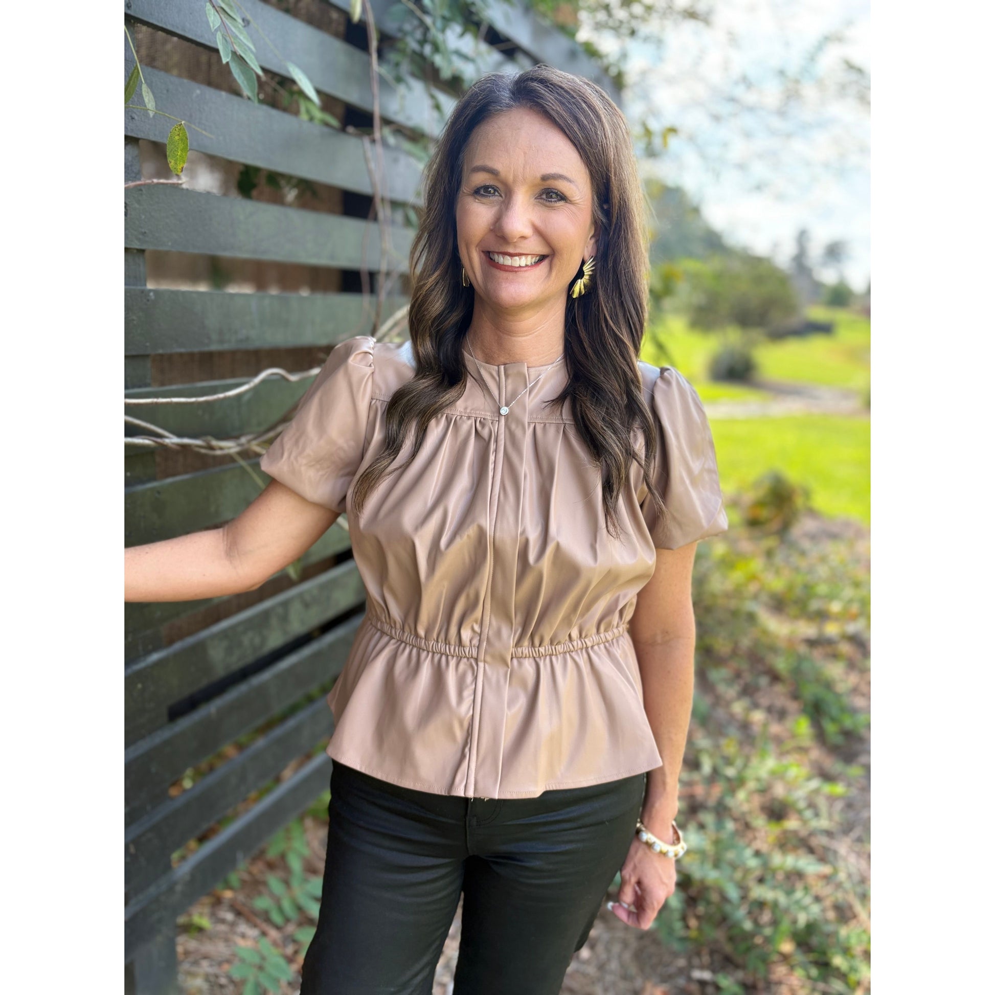 Woman wearing a beige blouse standing outdoors with greenery in the background