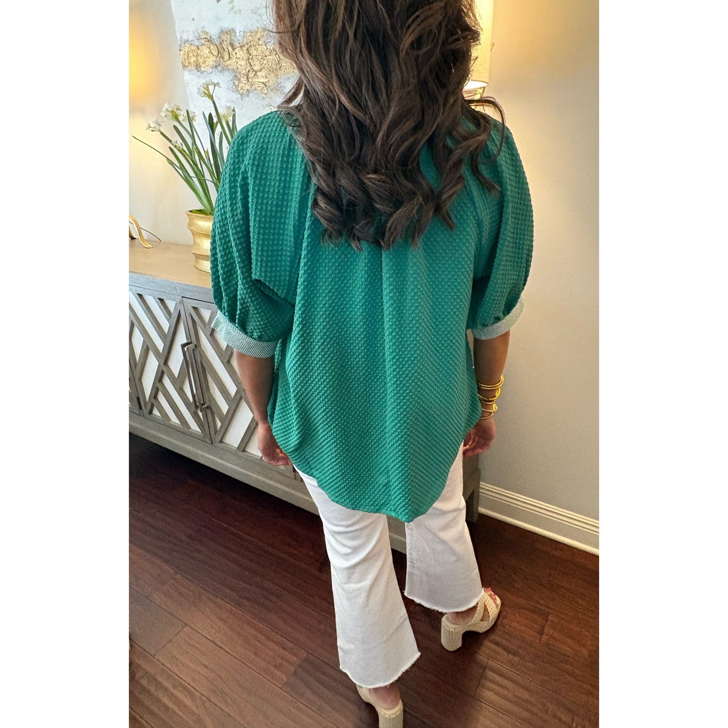 Person wearing a green sweater and white pants in a room with wooden flooring and a decorative cabinet.