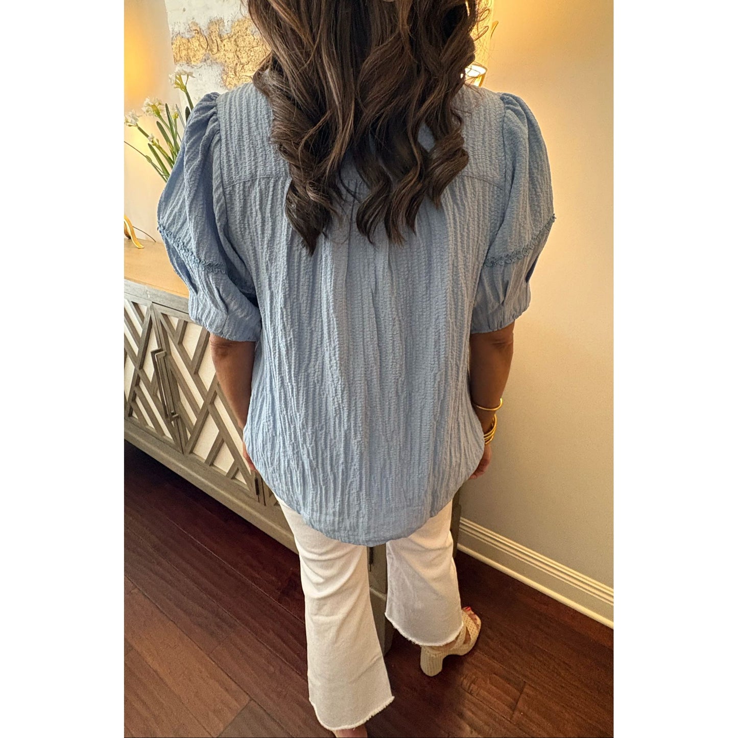 Person wearing a light blue textured blouse and white pants in a room with wooden flooring and decorative elements.