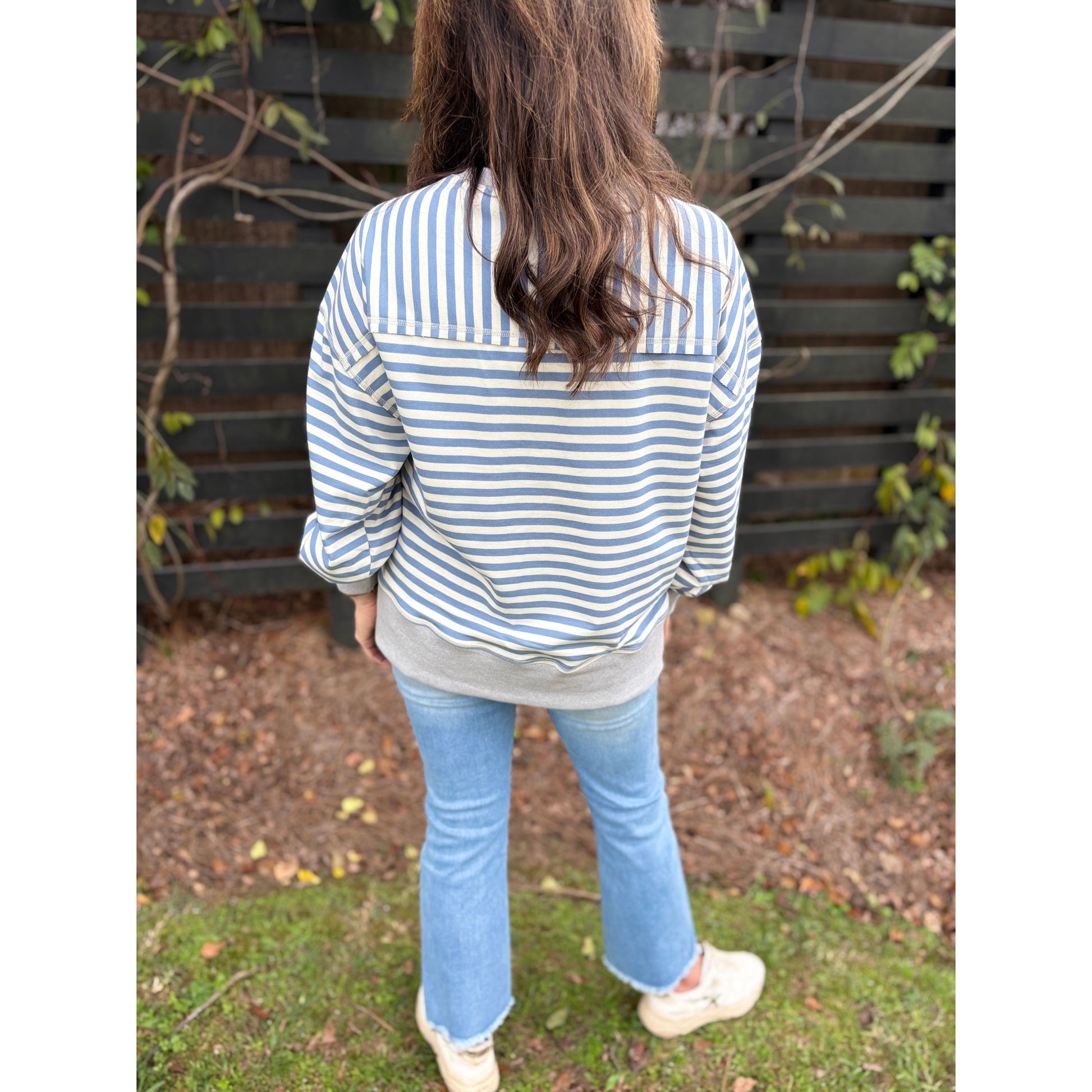 Person wearing a striped shirt and jeans standing outdoors with a wooden fence and plants in the background