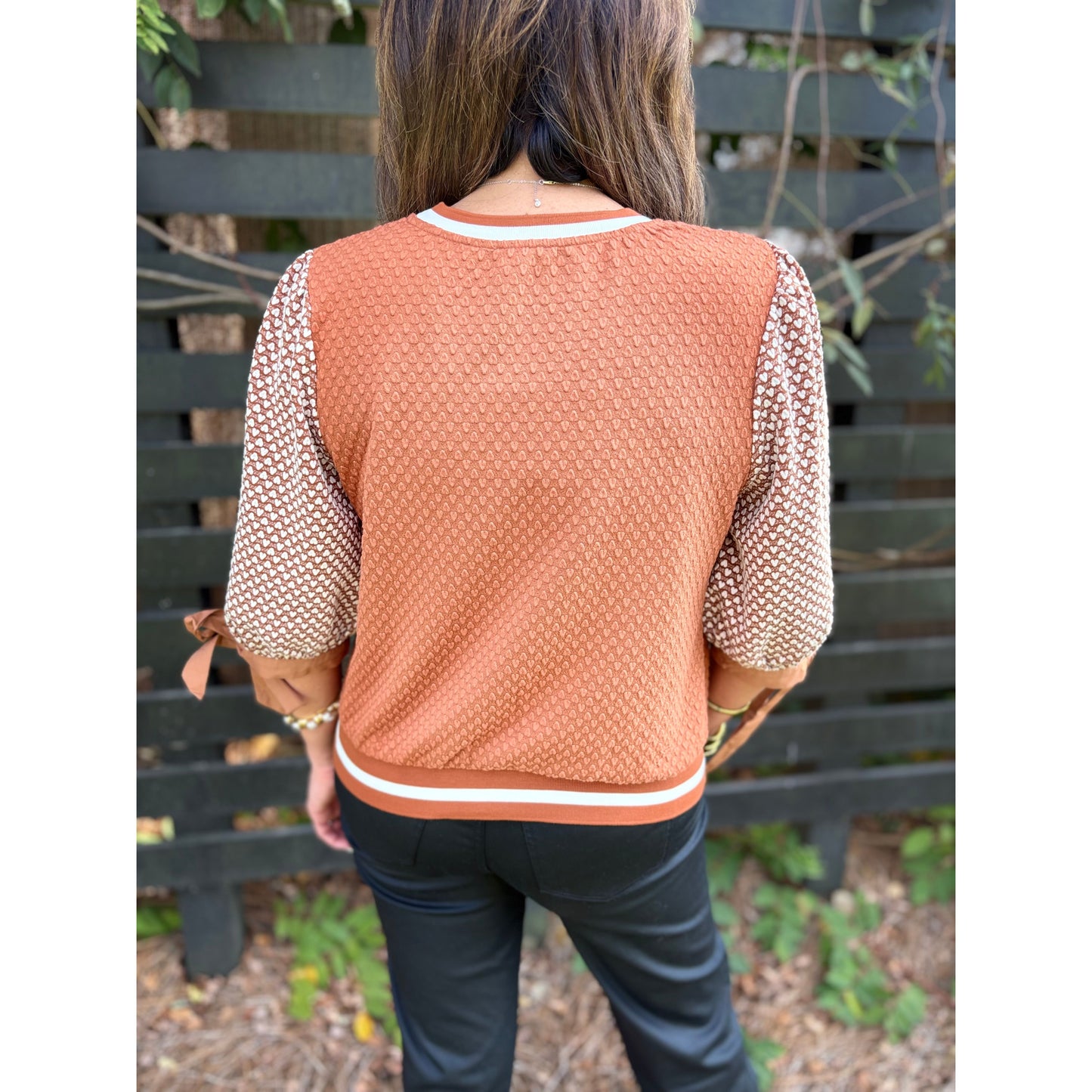 Person wearing a brown textured sweater with patterned sleeves against a wooden fence background