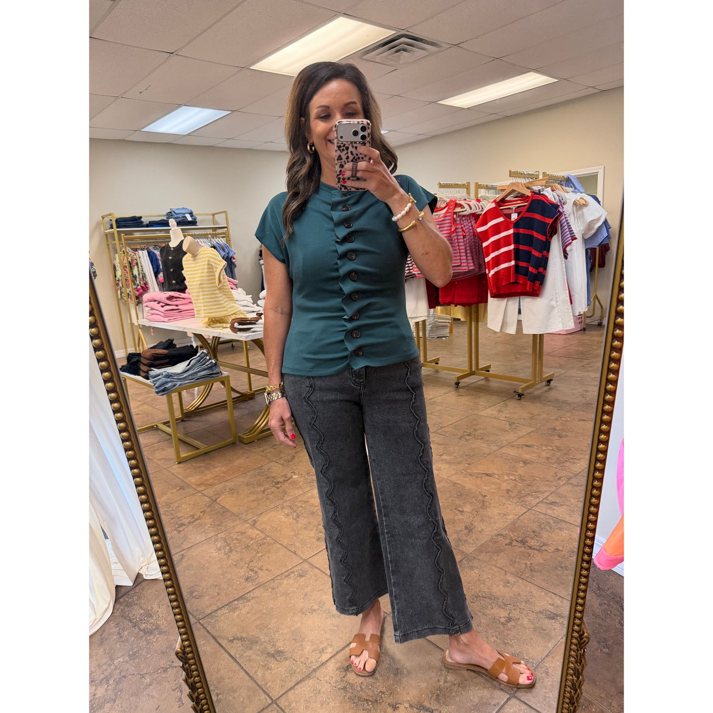 A woman takes a mirror selfie in a boutique wearing a teal short-sleeve top with a button-down front and dark gray wide-leg pants. Clothing racks and store fixtures are visible in the background.