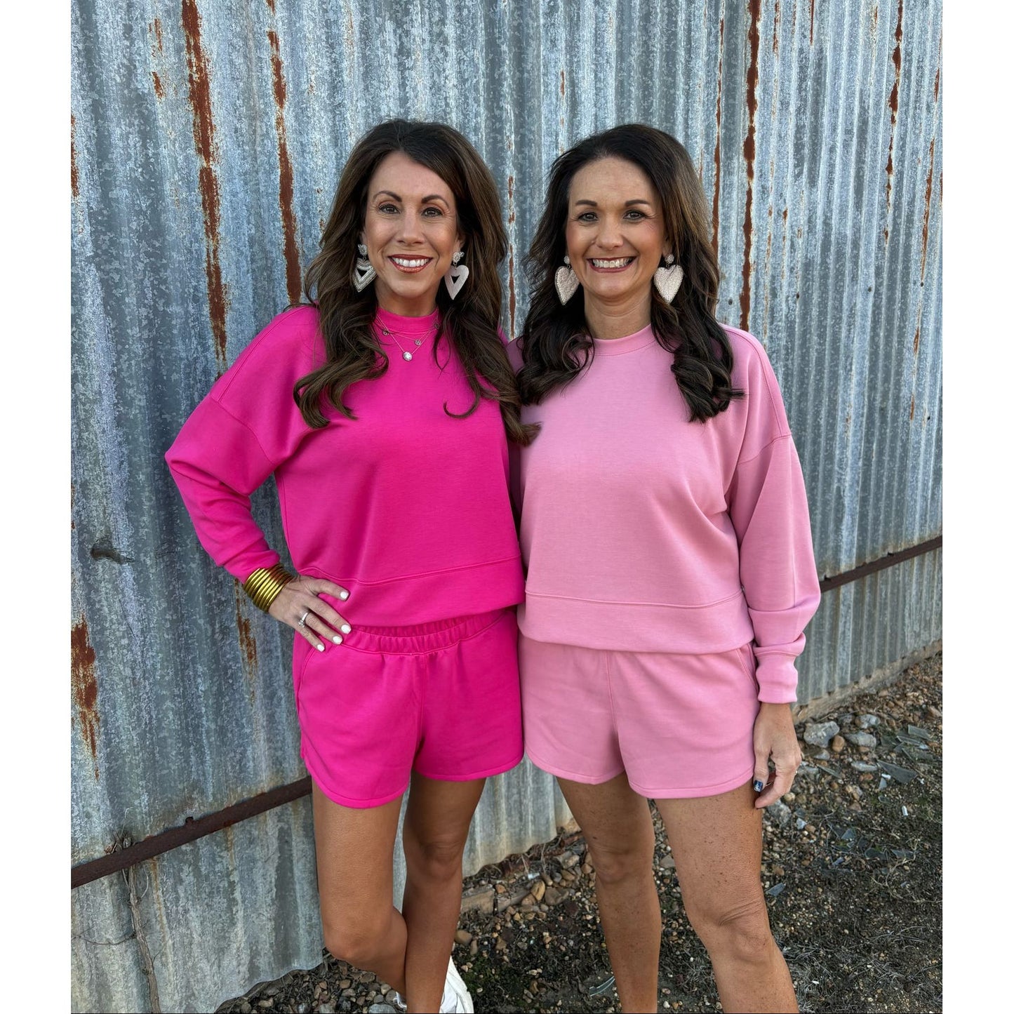 Two women in matching pink outfits standing against a corrugated metal wall.