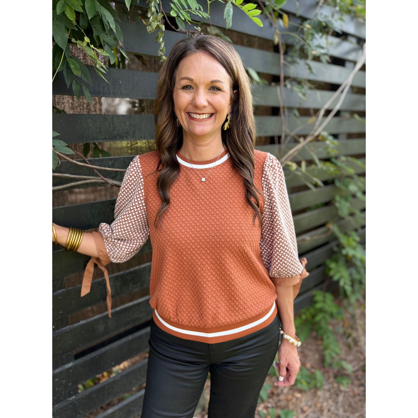 Woman wearing a brown patterned top and black pants standing against a wooden fence.