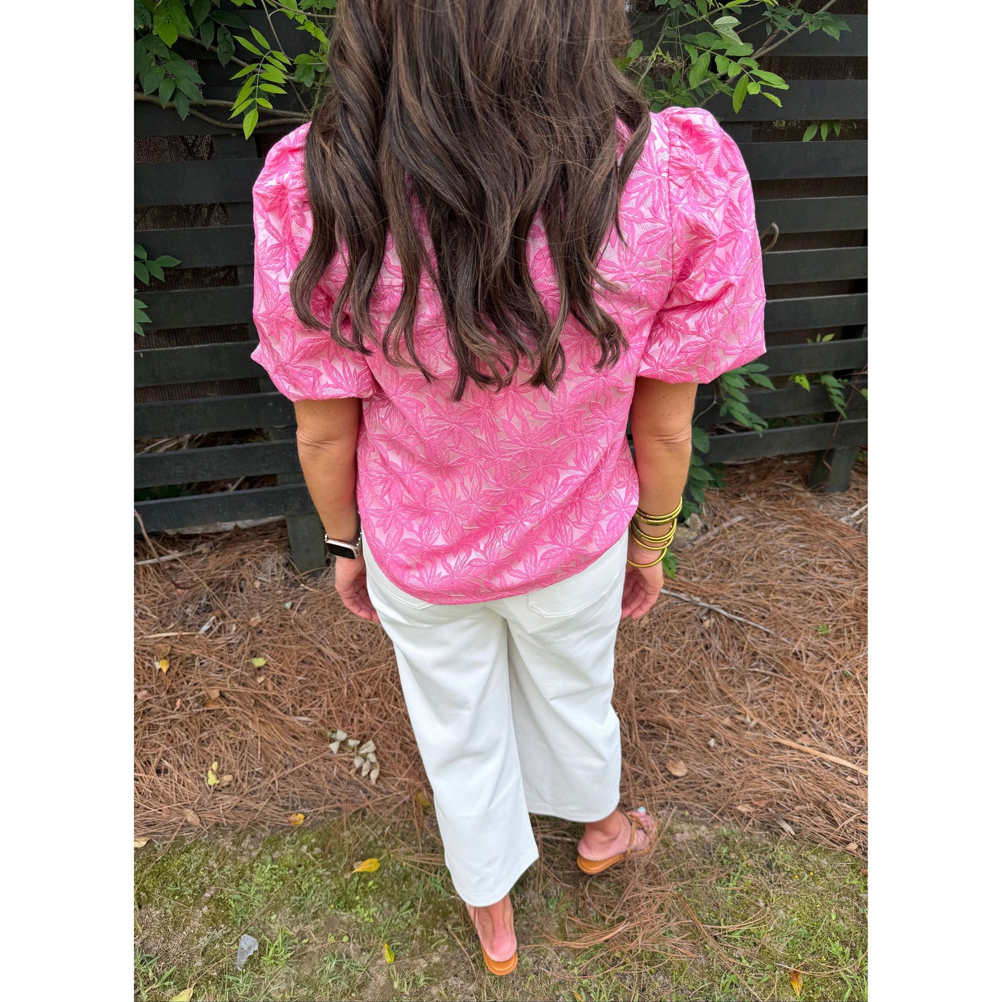 Person wearing a pink jacket and white pants standing on a grassy area with plants around.