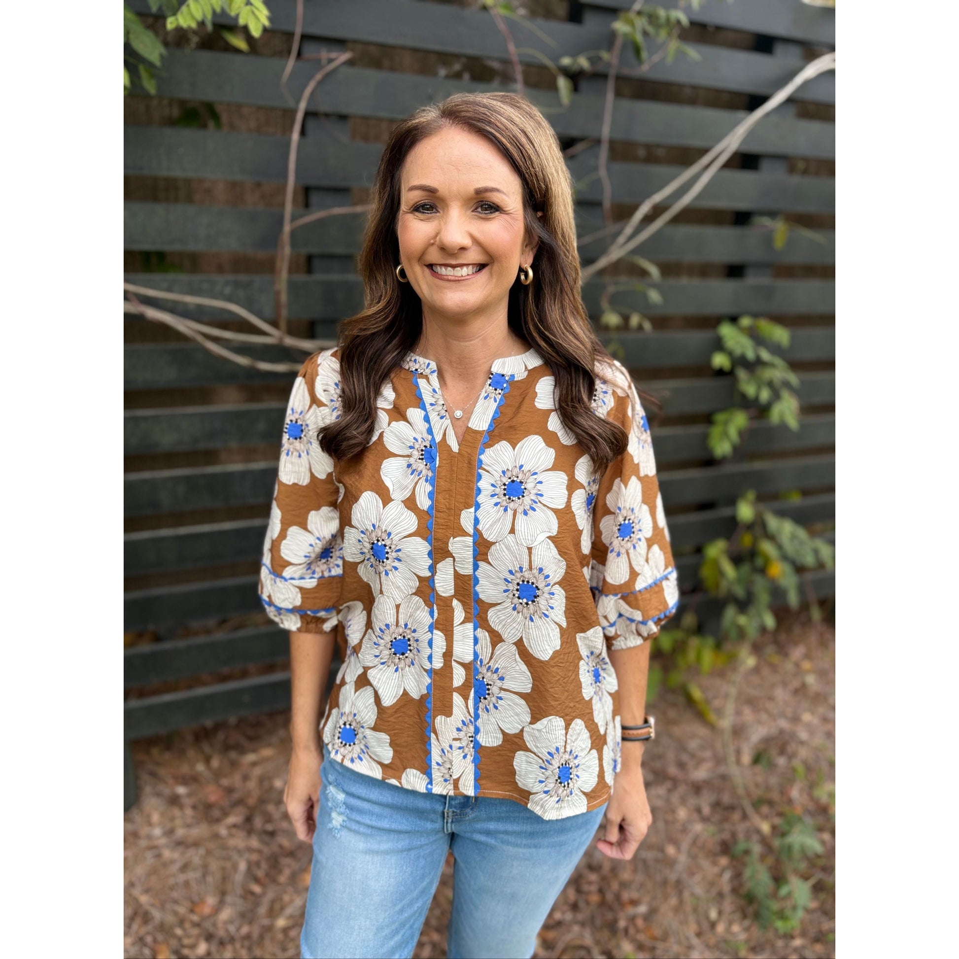 Woman wearing a floral blouse and jeans standing against a wooden fence.