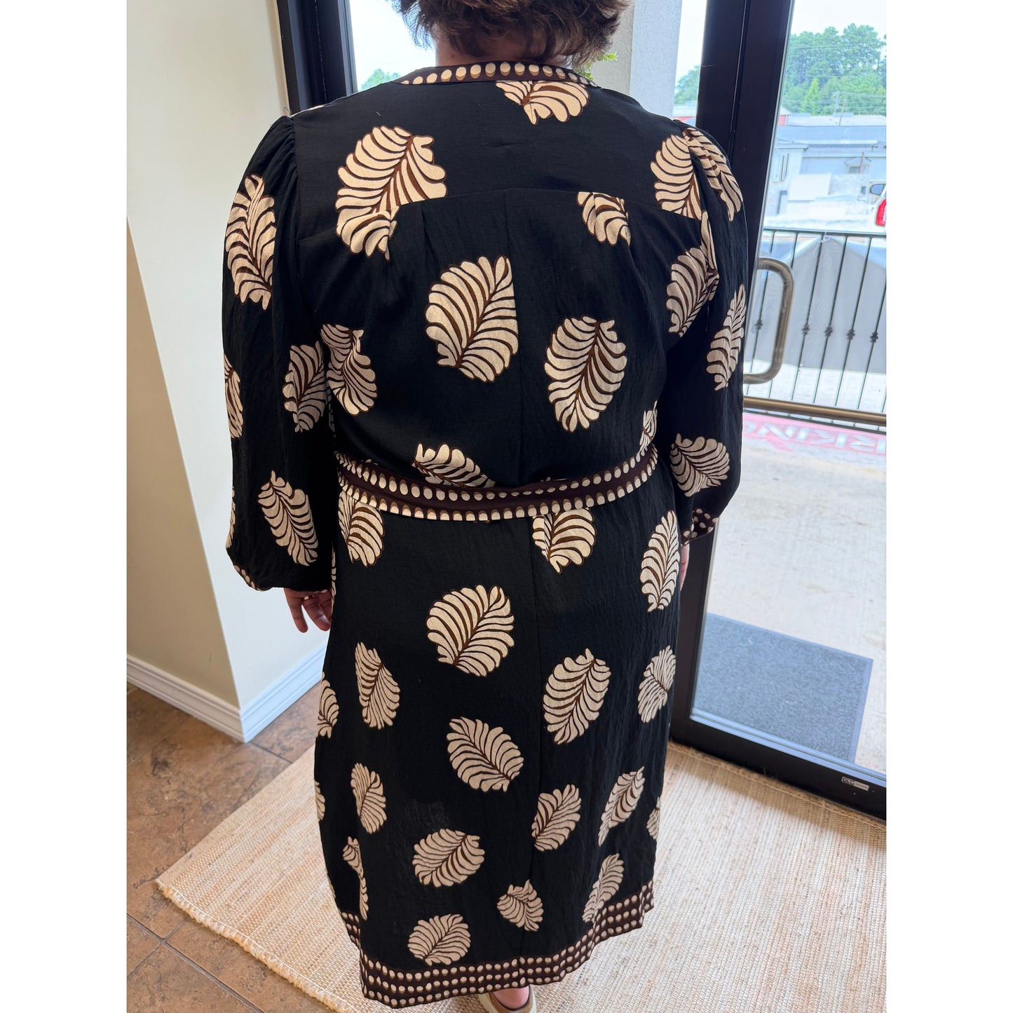 A woman viewed from behind wearing a black long-sleeve dress with a beige leaf pattern and a matching beaded belt, standing indoors near a glass door.