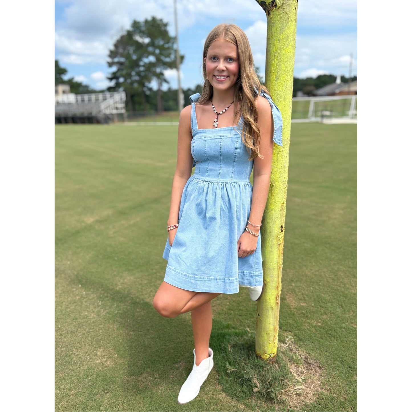 Young girl in a light blue dress standing next to a tree on a grassy field.