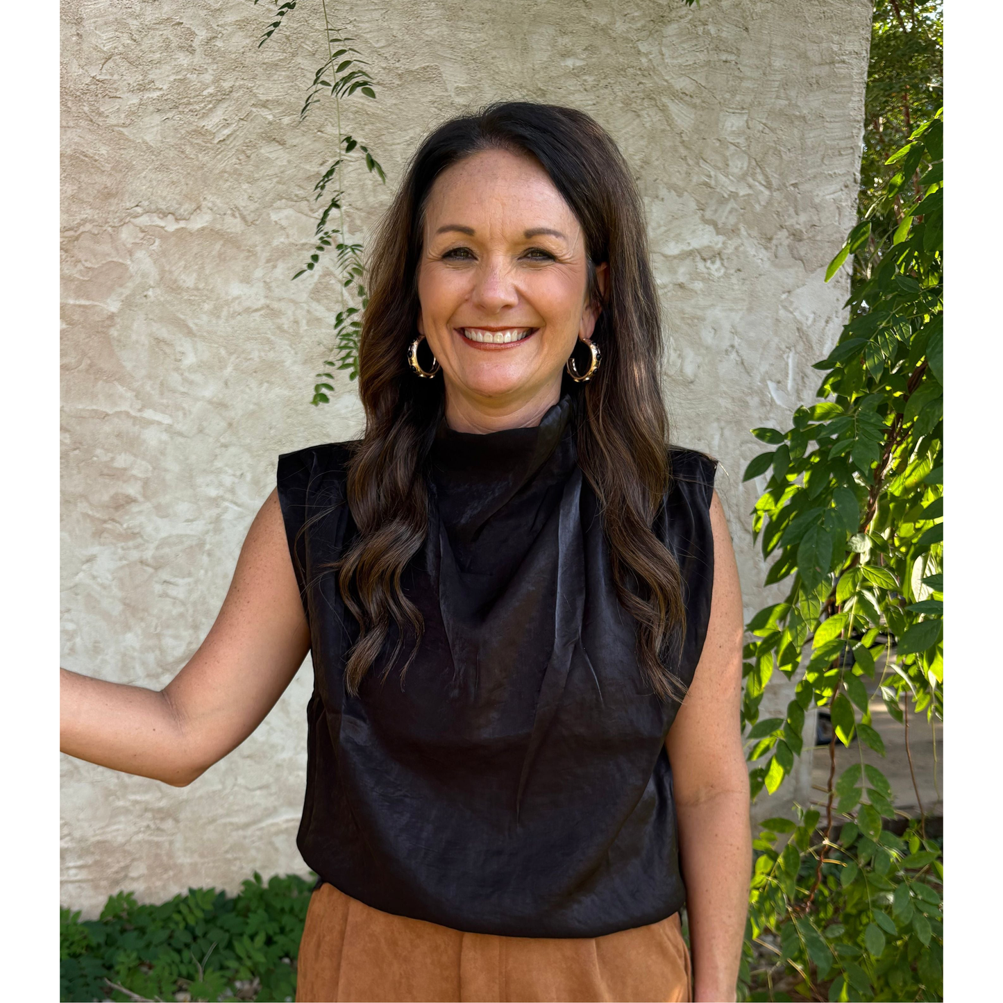 Woman wearing a black sleeveless top and brown pants standing against a stone wall with greenery.