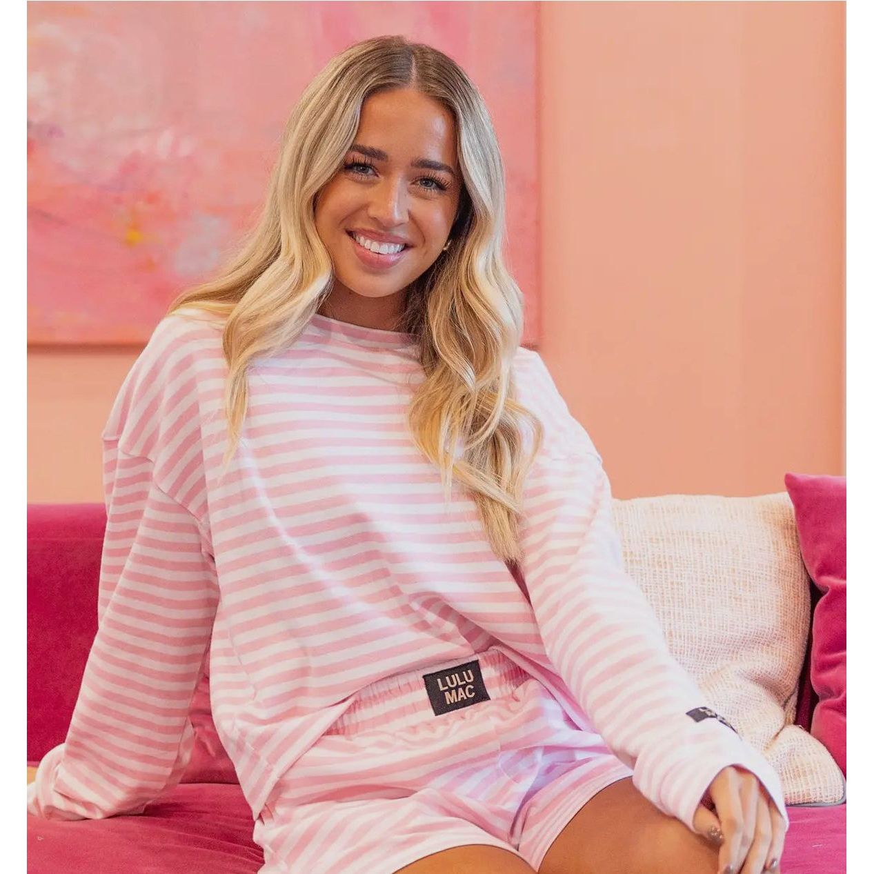 Woman wearing a pink striped outfit with 'Lulu' branding, sitting on a couch against a pink wall.