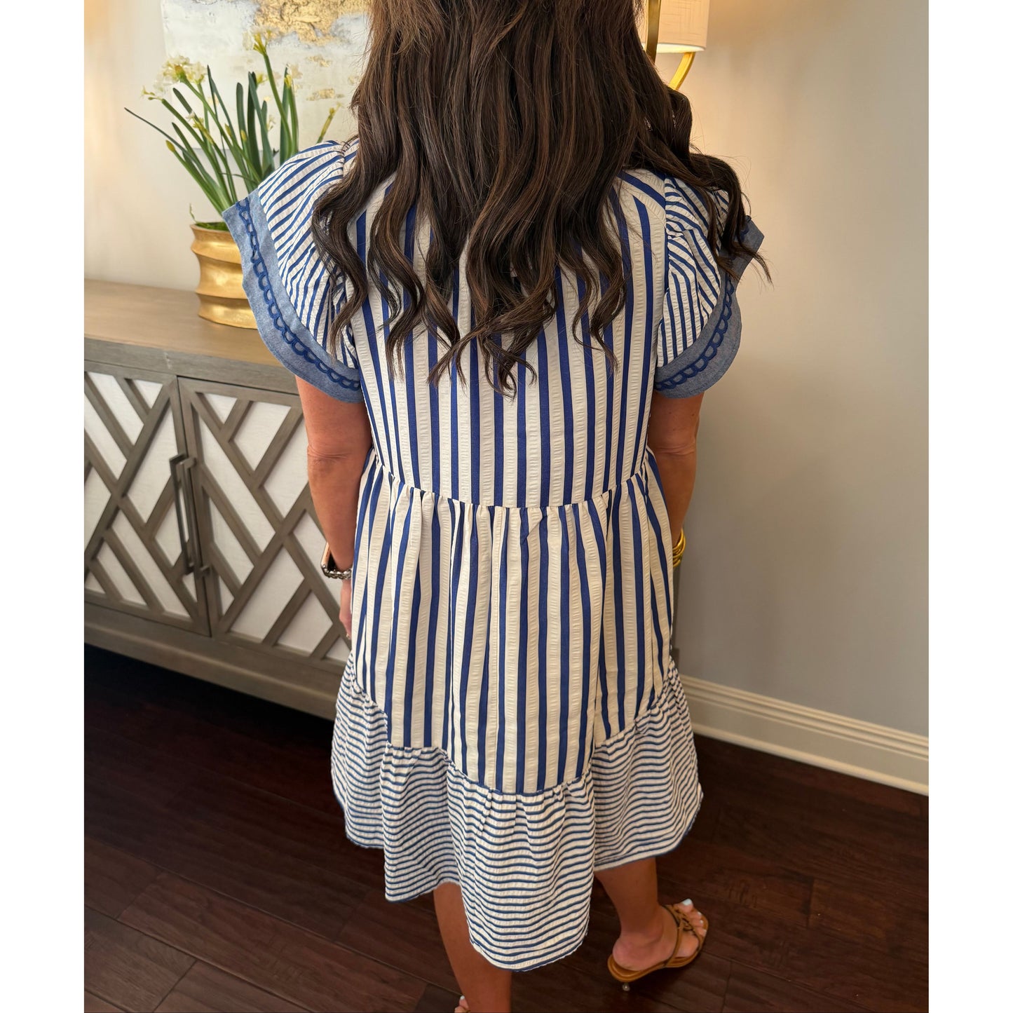 Person wearing a blue and white striped dress standing in a room with a lamp and decorative items.