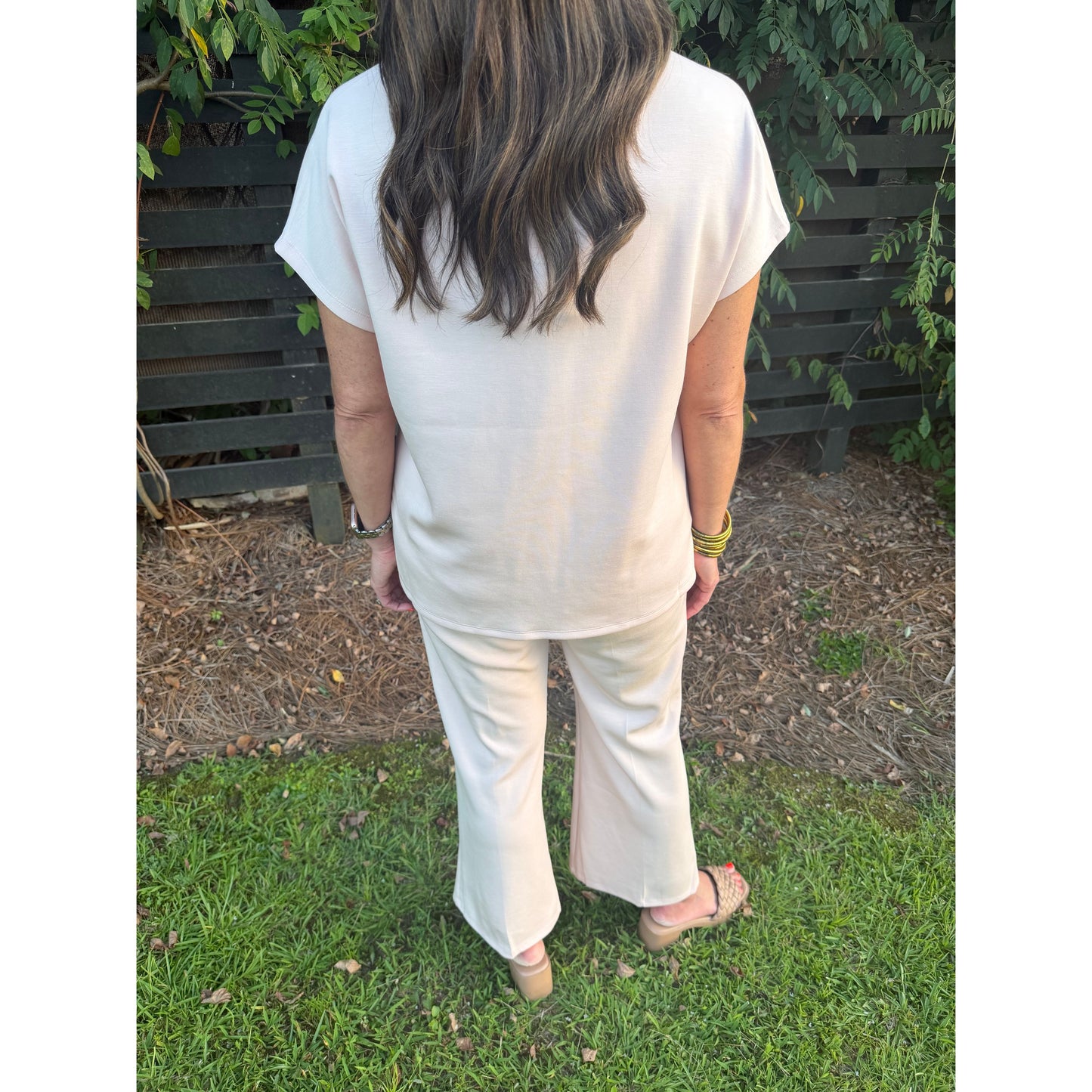A woman viewed from behind wearing a cream-colored short-sleeve top and matching wide-leg pants, standing outdoors on a grassy lawn with a dark wooden fence and greenery in the background.