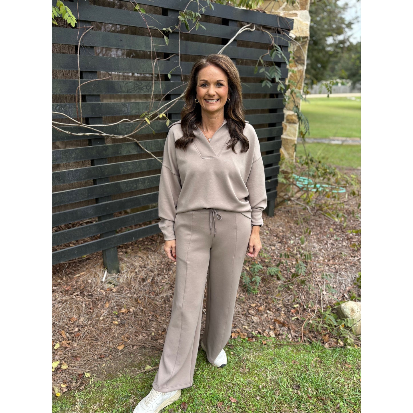 Woman in a beige tracksuit standing outdoors with a wooden fence and grass in the background