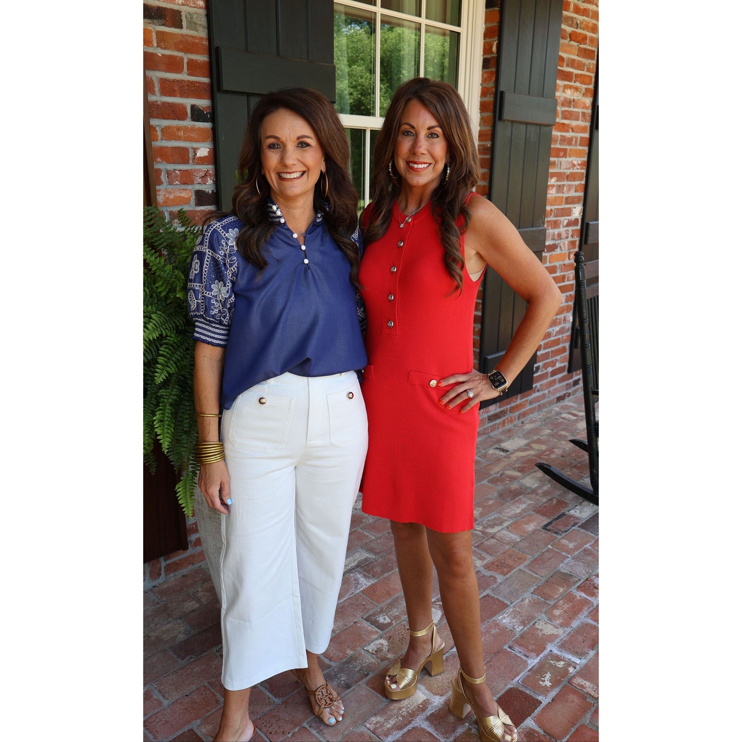 Two women standing on a brick porch, one in a blue top and white pants, the other in a red dress.