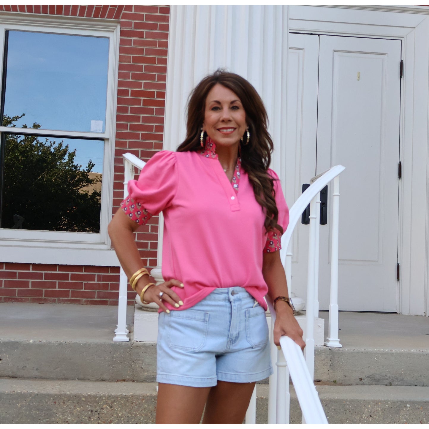 Woman in pink shirt and denim shorts standing on a brick building's steps.