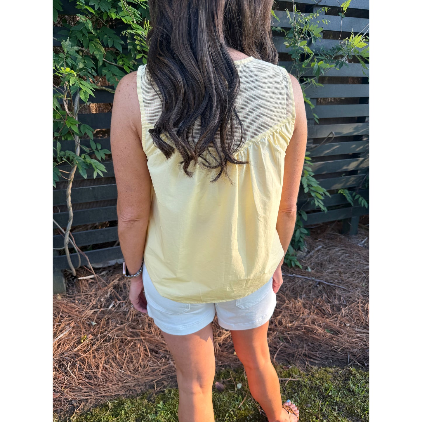 Person wearing a light yellow sleeveless top and white shorts standing outdoors with greenery in the background.