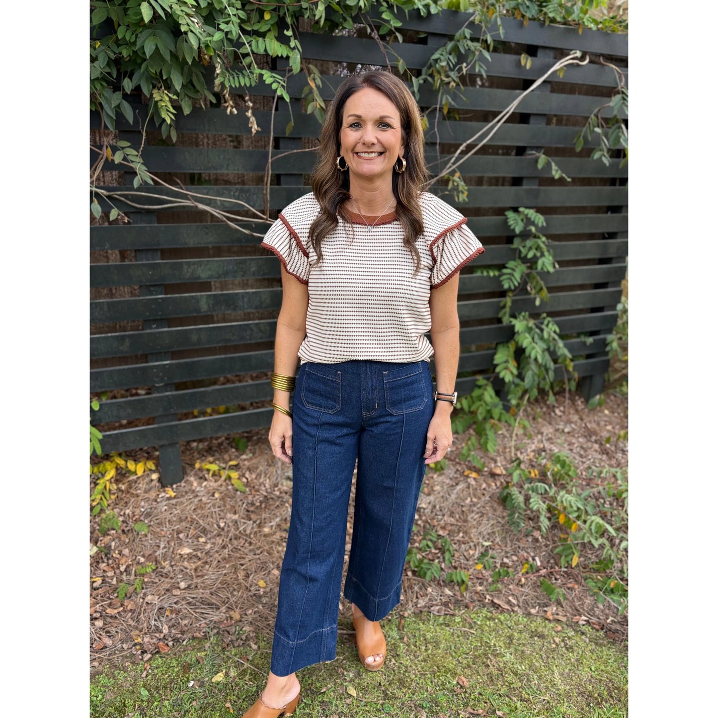 Woman wearing a striped shirt and blue jeans standing in front of a wooden fence.