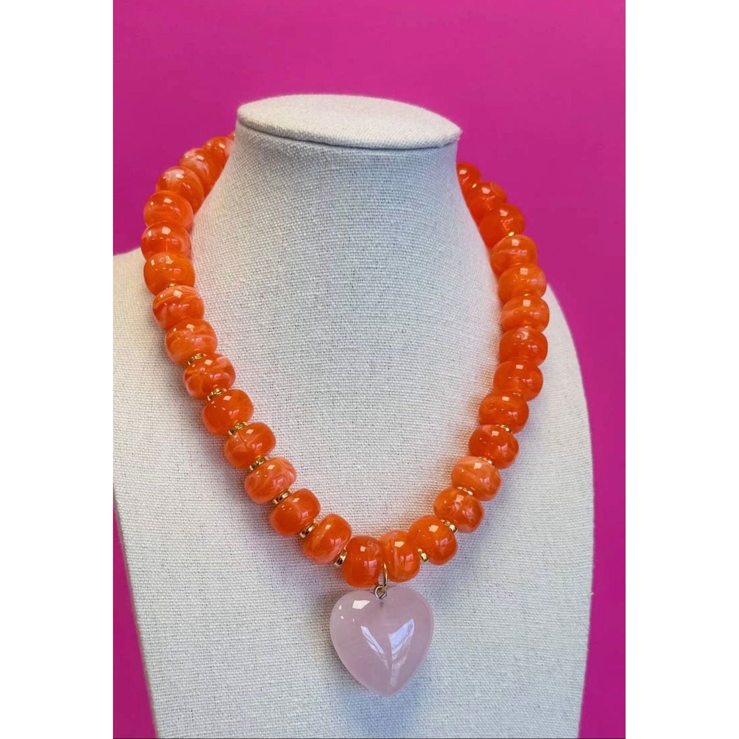 Necklace with orange beads and a pink heart pendant on a mannequin bust against a pink background