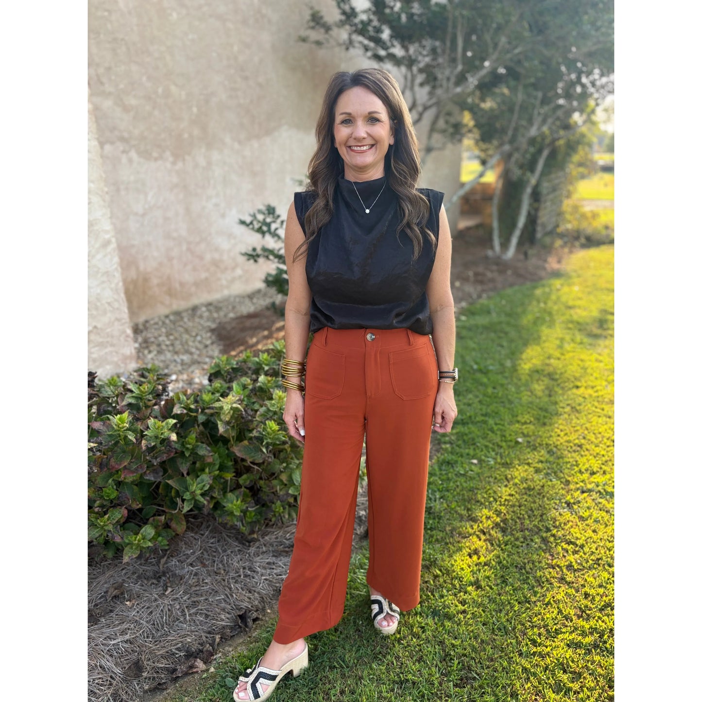 Woman wearing a black sleeveless top and rust-colored pants standing outdoors.