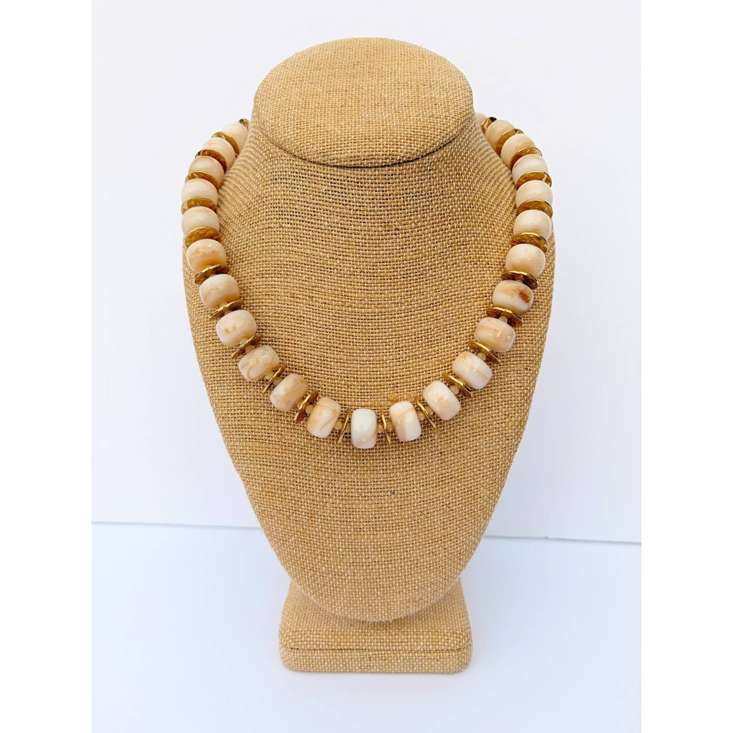 A necklace made of round cream-colored beads separated by small gold spacer rings, displayed on a tan fabric jewelry bust against a white background.