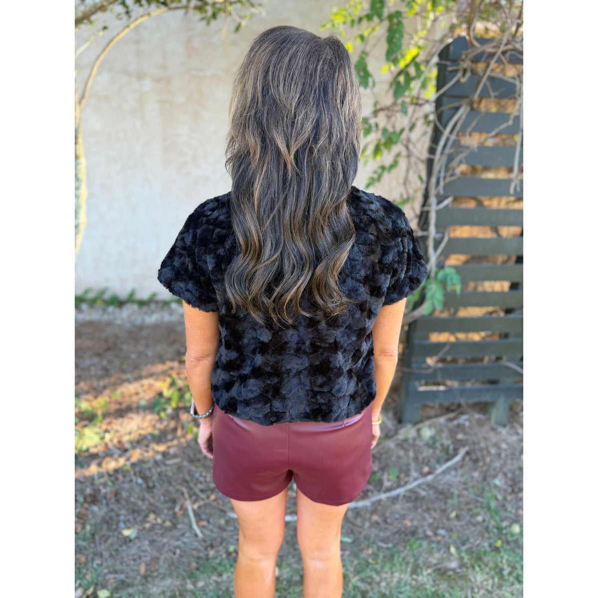 Annie Ks Boutique wine faux leather shorts with pintuck detail worn with a black textured top, back view