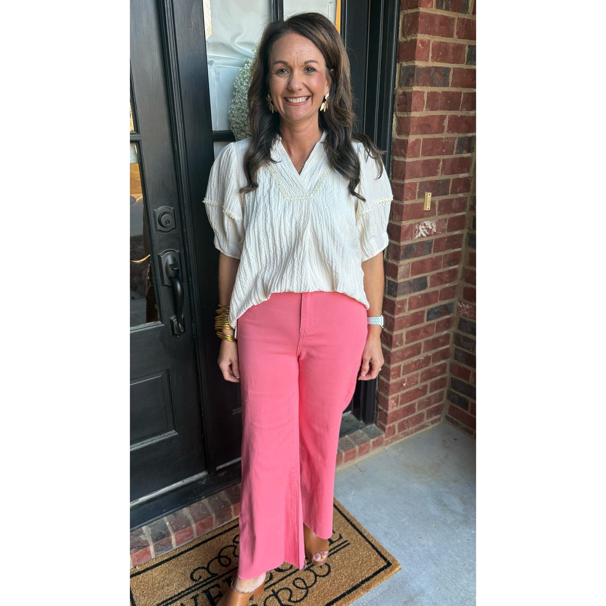 Woman in a white blouse and pink pants standing in front of a door.
