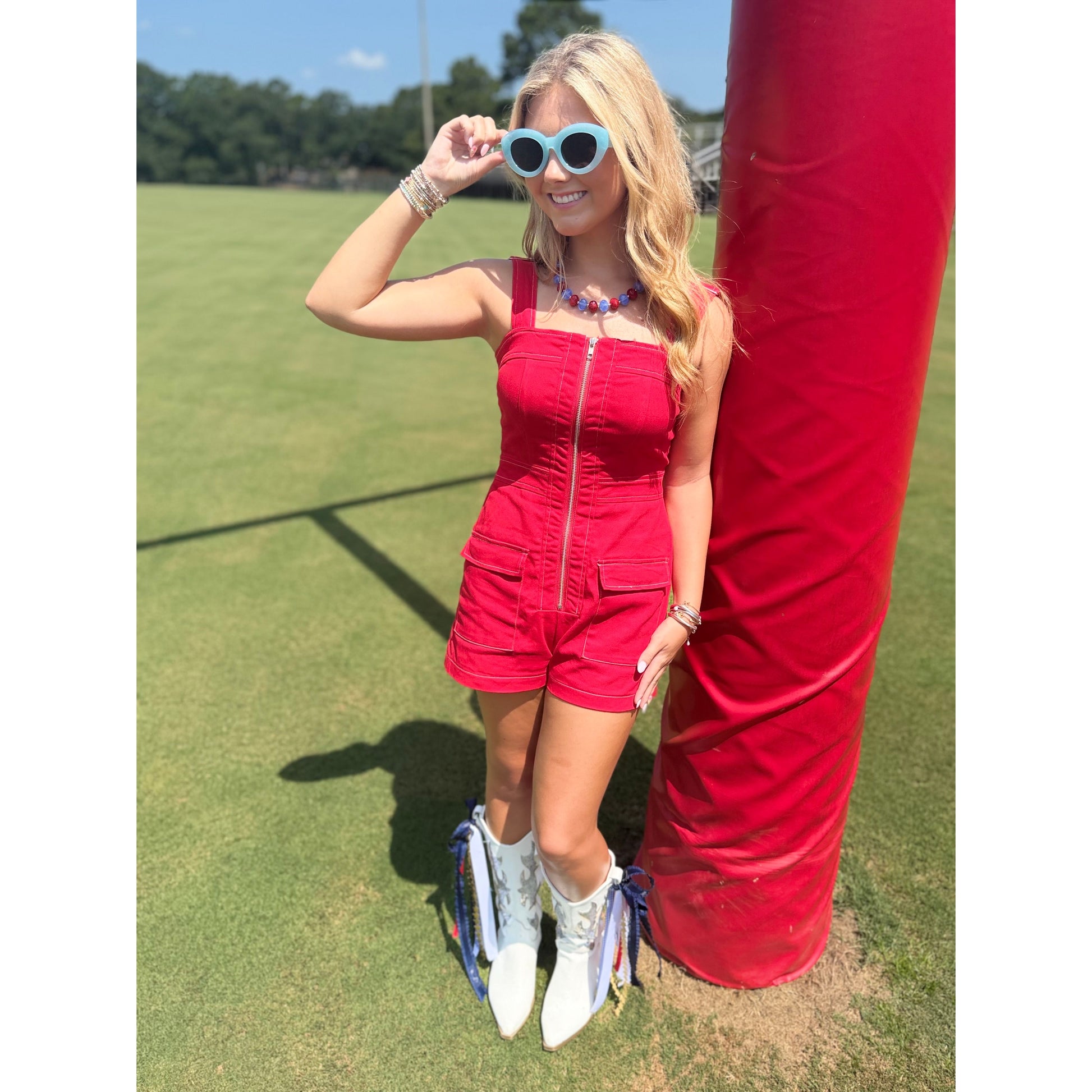 A woman stands outdoors on a grassy field next to a large red cylindrical pole, wearing a bright red sleeveless romper with a front zipper, white cowboy boots, and light blue heart-shaped sunglasses.