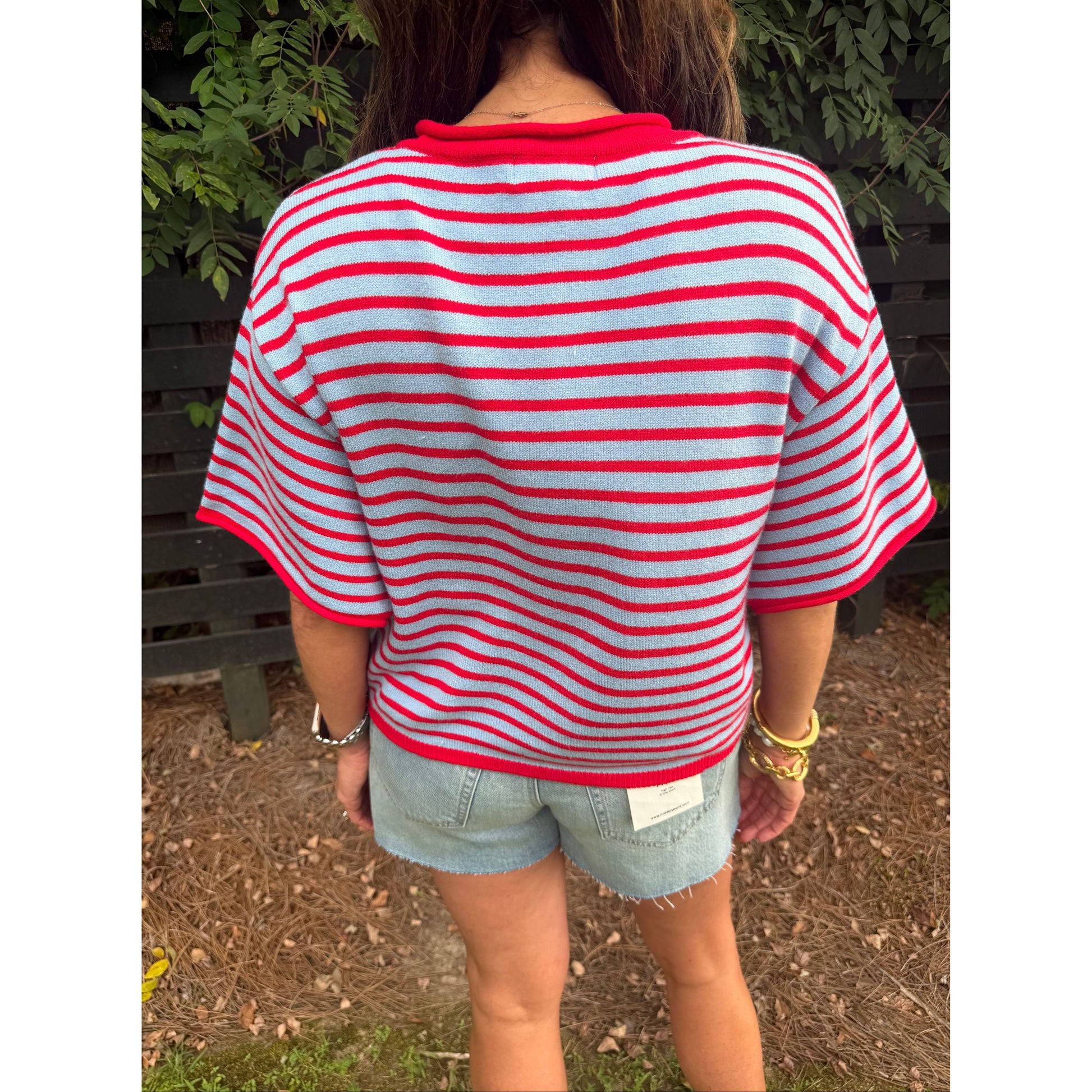 A person viewed from behind wearing a red and white striped short-sleeve knit top with a red crew neck, paired with denim shorts, standing outdoors on a mulch ground with green foliage in the background.