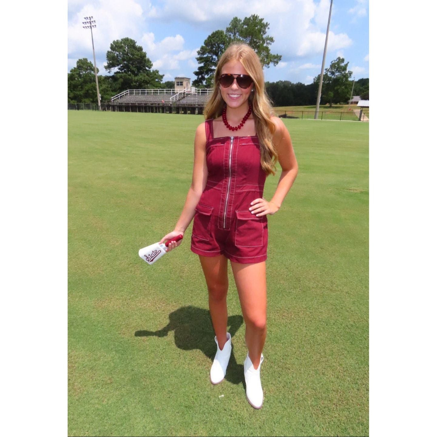 A woman stands on a grassy field wearing a sleeveless burgundy romper with a front zipper, holding a white spray bottle.