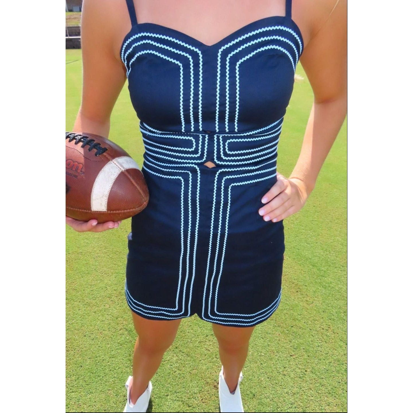 A woman wearing a navy blue sleeveless romper dress with white geometric piping, holding a brown football on a green grass field.