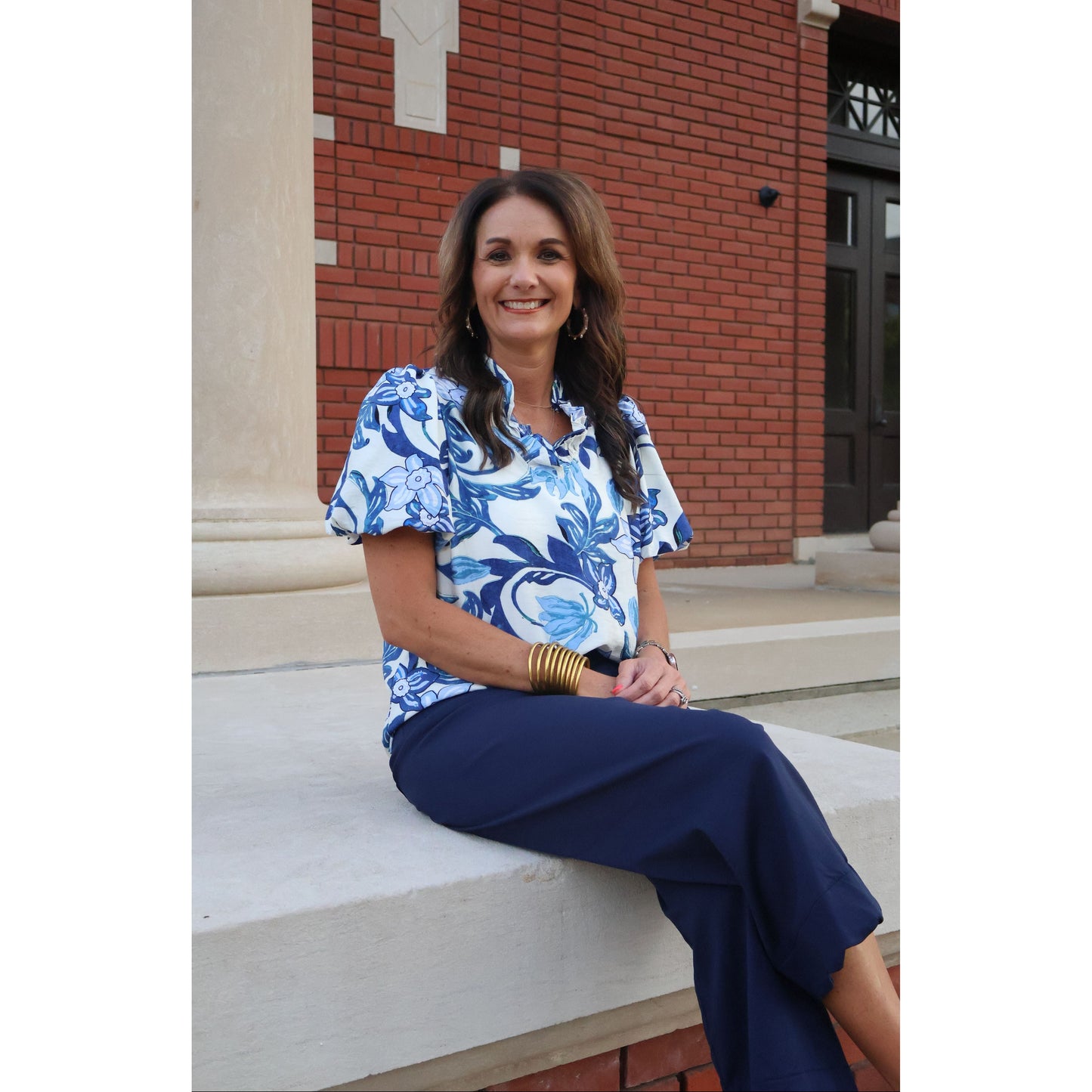Woman wearing a blue and white floral top with navy pants standing in front of a brick building.