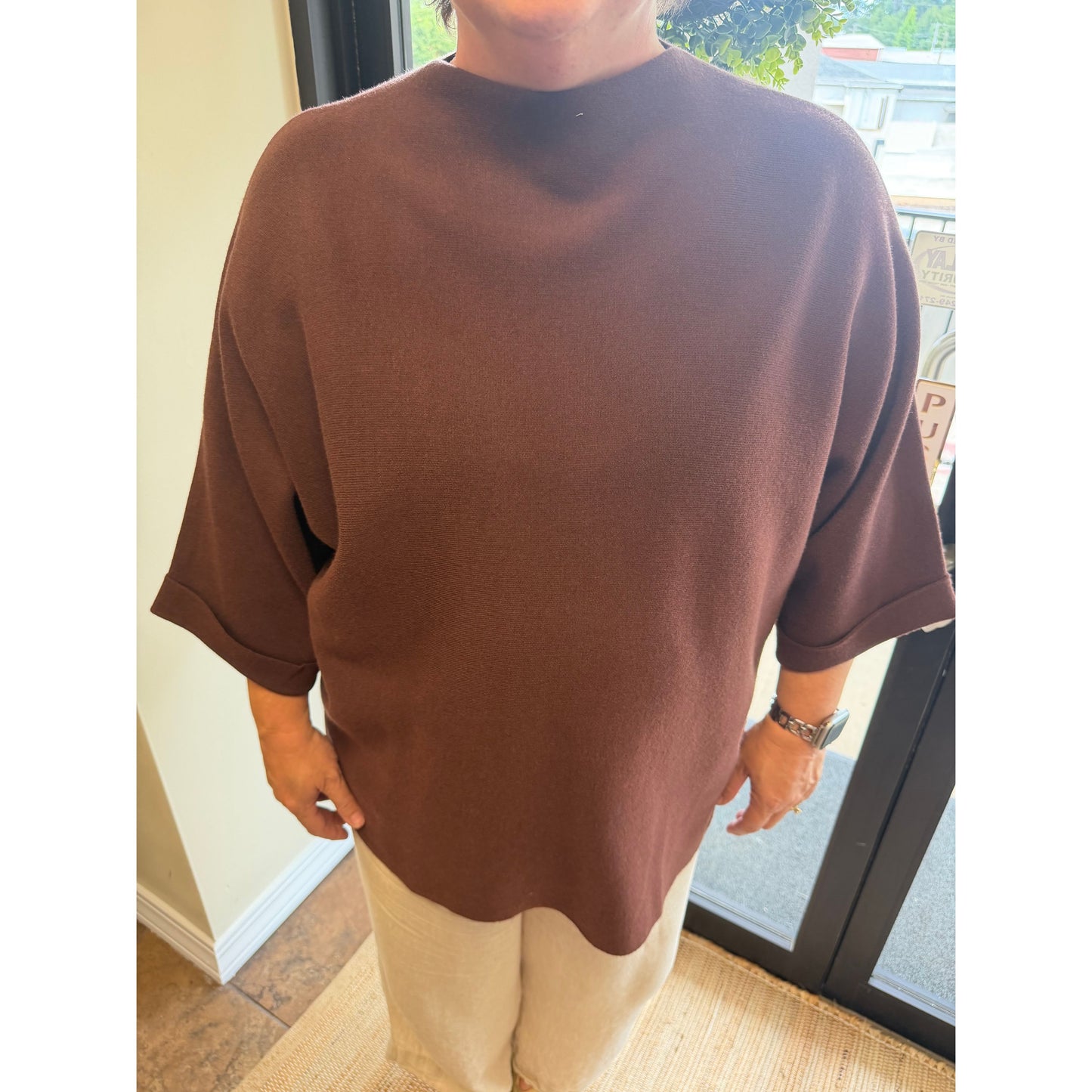 A person wearing an oversized short-sleeve knit top in a solid dark brown color, paired with light beige pants, standing indoors near a window.