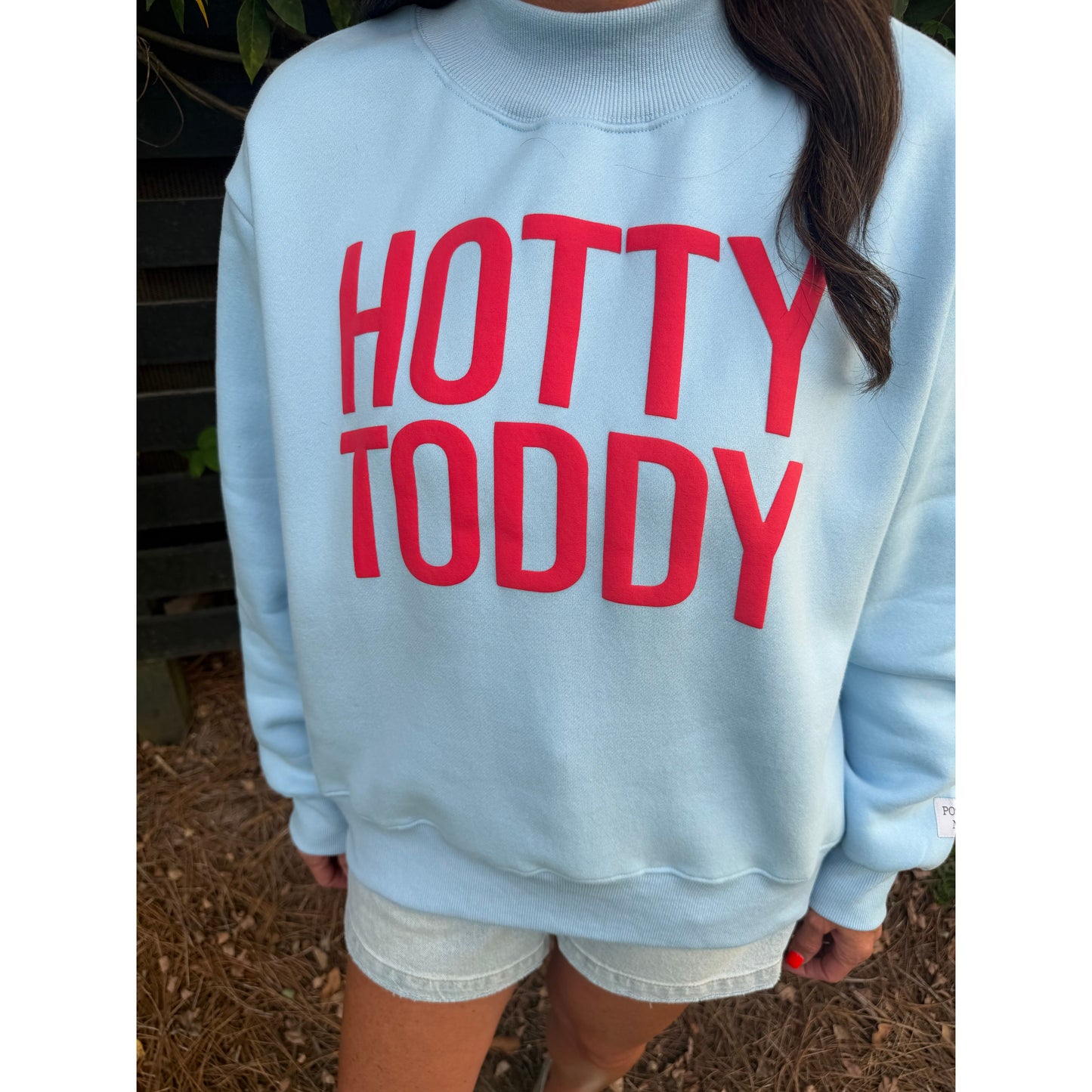 A person wearing a light blue sweatshirt with the words 'HOTTY TODDY' printed in large red letters on the back, paired with white denim shorts, photographed outdoors on a bench.