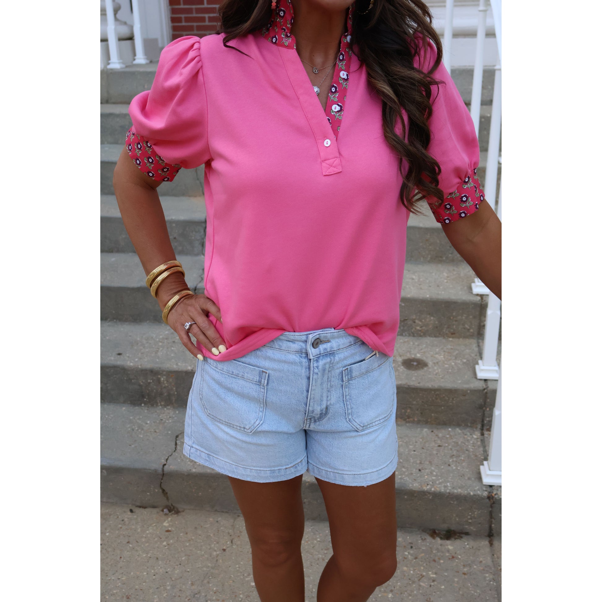 Person wearing a pink shirt with decorative elements and light blue shorts against a neutral background.