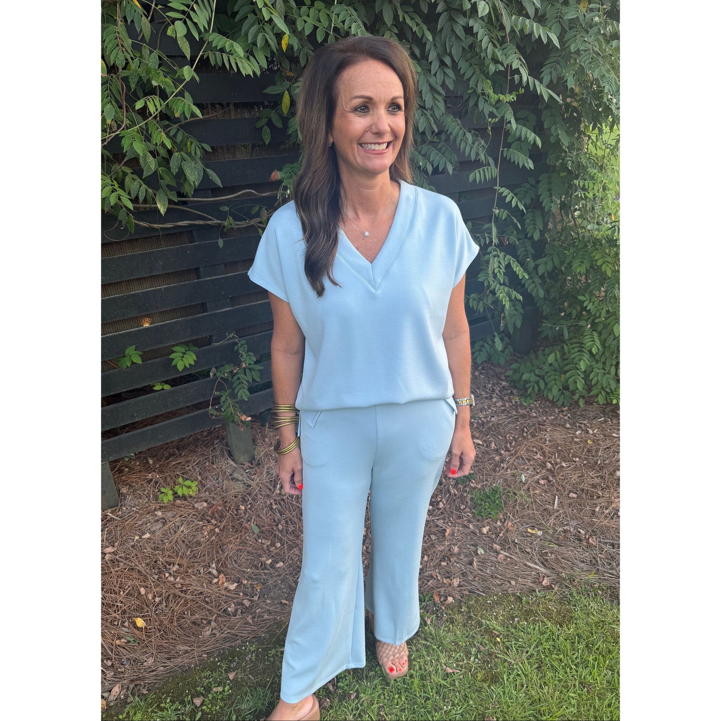 A woman wearing a light blue matching two-piece set consisting of a short-sleeve V-neck top and wide-leg pants, standing outdoors in front of a dark wooden fence and green foliage.