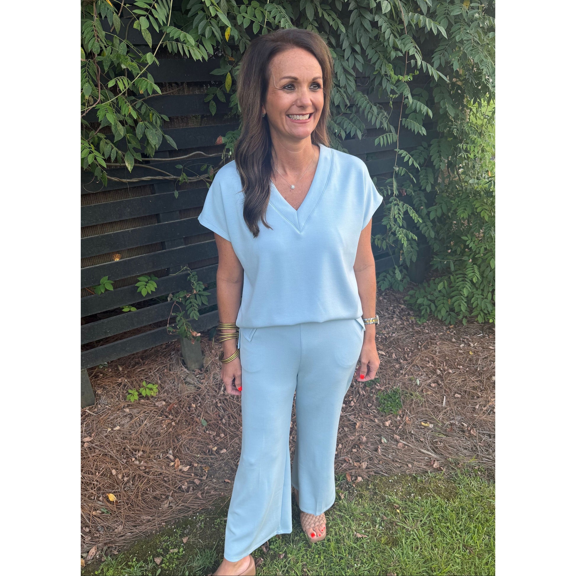 A woman wearing a light blue matching two-piece set consisting of a short-sleeve V-neck top and wide-leg pants, standing outdoors in front of a dark wooden fence and green foliage.