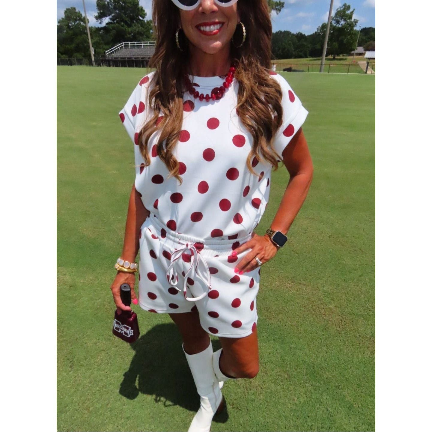 A woman stands on a grassy golf course wearing a white short-sleeve romper with red polka dots, white knee-high socks, and white boots. She is also wearing red beaded necklaces, hoop earrings, and sunglasses.